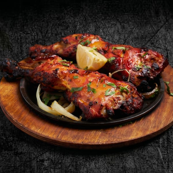 Sizzling tandoori chicken drumsticks with lemon and onions on a black plate and wooden board.