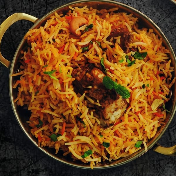 Golden biryani with chicken, cashews, and mint in a brass bowl on a dark textured surface.