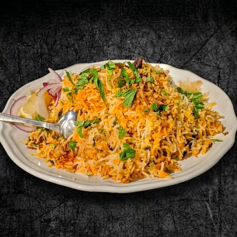 A white plate filled with colorful biryani rice, garnished with fresh mint, red onions, and lemon wedges.
