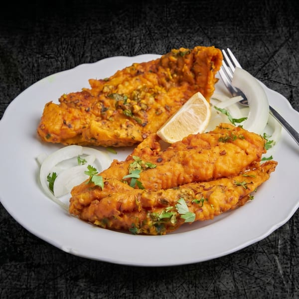 Two pieces of fried fish pakora with onion slices, cilantro, and a lemon wedge on a white plate.